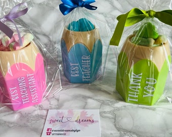 May include: Three pencil-shaped containers filled with colorful marshmallows. The containers are wrapped in clear plastic with a ribbon. The containers are labeled "Best Teaching Assistant", "Best Teacher", and "Thank You".