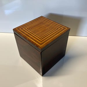 May include: A small, square wooden box with a dark brown exterior and a light brown geometric pattern on the lid.