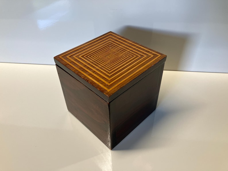 May include: A small, square wooden box with a dark brown exterior and a light brown geometric pattern on the lid.