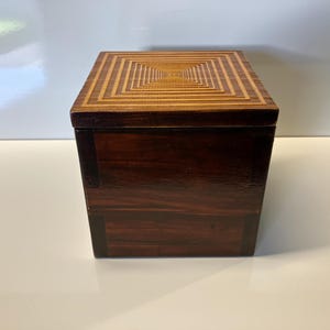 May include: A wooden box with a geometric pattern on the lid. The box is made of dark wood with a light wood inlay.