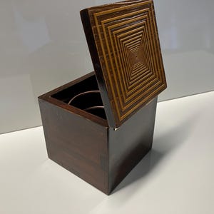 May include: A wooden box with a hinged lid. The lid has a geometric pattern of concentric squares in shades of brown. The box is closed and sitting on a white surface.