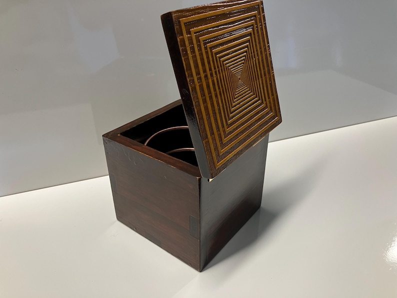 May include: A wooden box with a hinged lid. The lid has a geometric pattern of concentric squares in shades of brown. The box is closed and sitting on a white surface.