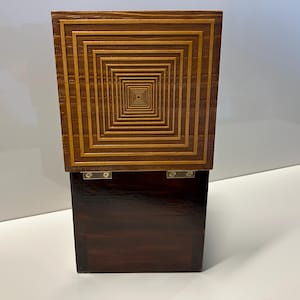 May include: A wooden box with a dark brown base and a top made of light wood with a geometric pattern of concentric squares.