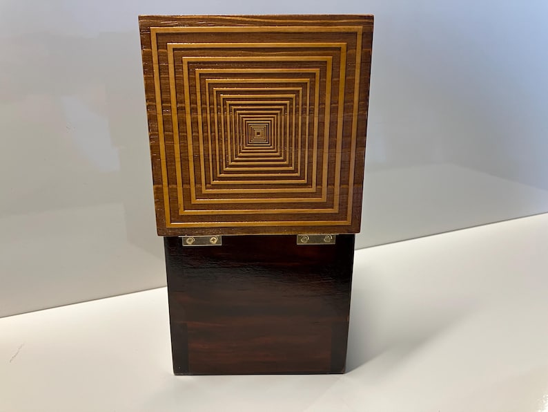 May include: A wooden box with a dark brown base and a top made of light wood with a geometric pattern of concentric squares.
