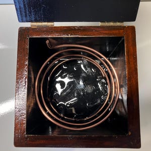 May include: A wooden box with a copper coil inside. The coil is submerged in a black liquid.