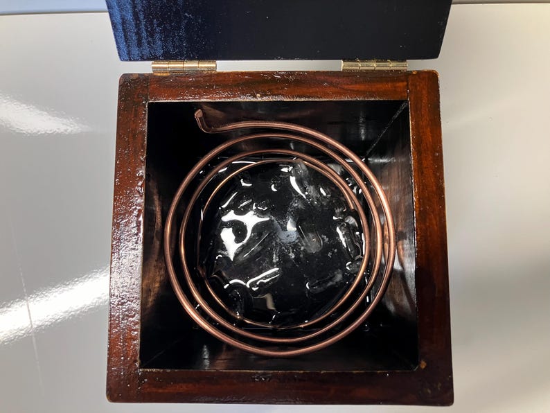 May include: A wooden box with a copper coil inside. The coil is submerged in a black liquid.