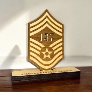 May include: A wooden plaque with a brown and white chevron design, a star, and the number 137. The plaque is on a wooden base with the text "CMSgt. Jonhcek 11/2023".