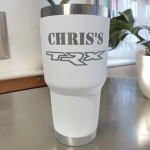 May include: White stainless steel tumbler with a silver lid. The tumbler has the text "CHRIS'S TRX" printed on the side in a gray font.