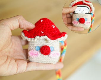 May include: A handmade crochet AirPod case shaped like a mushroom. The mushroom has a red cap with white dots, a beige body, and a face with pink cheeks, a red nose, and blue eyes. A rainbow strap is attached.