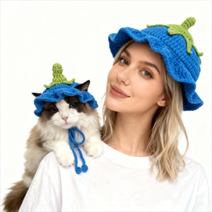 May include: A woman and a cat wearing matching blue crochet flower hats with green accents. The hats have a ruffled brim and a stem-like detail on top. The cat is light brown and white.