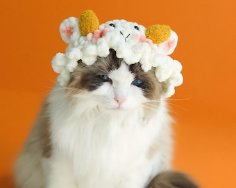 May include: A fluffy cat with blue eyes wearing a white and yellow crocheted lamb hat. The cat is sitting against an orange background. The hat has yellow ears and pink accents.