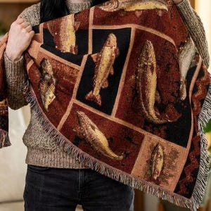 Peut inclure: Une couverture rectangulaire avec un motif de poissons répété en or, marron et noir. La couverture a une bordure à franges. La couverture est tenue par une personne. La couverture est faite d'une matière douce.