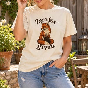 May include: A cream-colored short-sleeve t-shirt featuring a graphic of a sitting fox in the center. Above the fox, the text reads Zero, and below the fox, the text reads fox given in a dark brown, curved, bold, serif font.