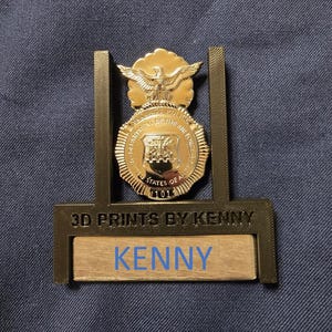May include: A gold-coloured 3D printed stand with a gold-coloured badge featuring an eagle and the text "Department of the Air Force" and "United States of America". The stand has a nameplate with the text "3D Prints by Kenny" and "Kenny".