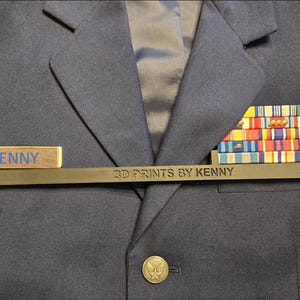 May include: A black metal name tag with the text "3D Prints by Kenny" and a wooden name tag with the text "KENNY". The name tags are attached to a blue uniform jacket with a row of colorful ribbons.