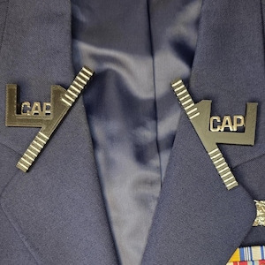 May include: Two black metal pins, each with the letters "CAP", are pinned to a navy blue blazer. The pins feature a ruler-like design. The blazer also has a ribbon and a small emblem. The pins are attached to the lapel of the jacket.