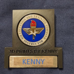 May include: A gold-colored 3D printed plaque with a blue and gold Air Education and Training Command Instructor emblem. The plaque has a slot for a nameplate, which reads "KENNY". The plaque is labeled "3D PRINTS BY KENNY".