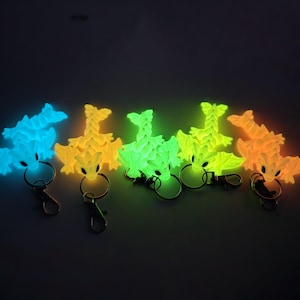 Może przedstawiać: Pięć świecących w ciemności breloków w kształcie smoka w różnych kolorach: niebieskim, żółtym, zielonym, jasnozielonym i pomarańczowym. Każdy brelok ma srebrny pierścień.