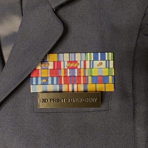 May include: A navy blue uniform jacket with a collection of colourful ribbons and a rectangular nameplate. The nameplate reads "3D PRINTS BY KENNY". The ribbons are in various colours including red, blue, yellow, and green.