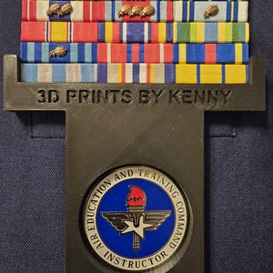 May include: A display of military ribbons with gold-colored embellishments, mounted on a black holder. The holder has the text "3D PRINTS BY KENNY" and a circular emblem with the text "AIR EDUCATION AND TRAINING COMMAND INSTRUCTOR".