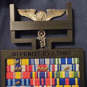 May include: A black 3D printed display stand with the text "3D PRINTS BY KENNY" on the front. The stand holds a silver pilot wings badge, a silver medical badge, and a collection of colourful military ribbons.