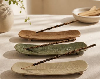 May include: Three elongated, curved ceramic incense holders with small feet, each featuring a unique leaf-like texture. The holders are coloured in shades of tan, sage green, and light beige, and each supports a single dark brown incense stick.