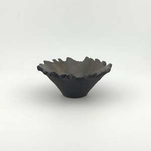 May include: A small, dark gray bowl with a rough, textured exterior and a wide, uneven, jagged rim. The interior surface of the bowl is smooth and dark, contrasting with the coarse, granular appearance of the outer walls and the irregular, organic shape of the top edge.