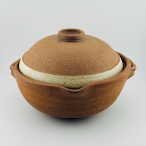 May include: A round, brown clay cooking pot with a matching domed lid. The pot features two small side handles and a textured, unglazed exterior. A wide, off-white band encircles the base of the lid, which is topped with a small, circular knob.