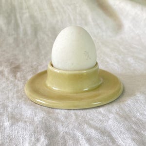 Puede incluir: Un huevo blanco en un huevera de cerámica amarillo pálido. La huevera tiene una base circular y un pequeño soporte redondeado. El fondo es una tela clara y texturizada.