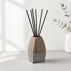 May include: A ceramic reed diffuser featuring a two-toned design with a matte tan upper section and a textured grey lower section. The vessel has a distinct vertical ridge along the front and contains seven thin black reeds extending upwards.