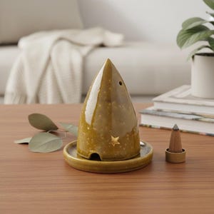 Może przedstawiać: Stożkowate, musztardowo-żółte ceramiczne kadzidełko z błyszczącym wykończeniem, posiadające małe wycięcie w kształcie gwiazdy przy podstawie oraz okrągły otwór. Stoi na pasującej okrągłej podstawce, obok znajduje się brązowy stożek kadzidlany.