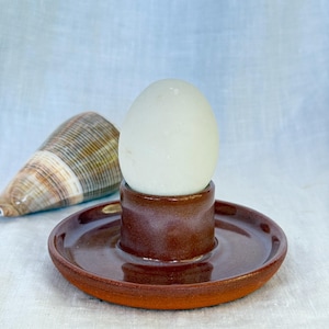 May include: A brown ceramic egg cup with a circular base, holding a white egg. The egg cup has a cylindrical center. A seashell is in the background. The egg cup is likely handmade.
