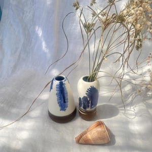 May include: Two ceramic vases with blue abstract designs and brown bases, alongside a seashell. One vase is taller, holding dried plants. The other vase is shorter. The seashell is light brown with a textured surface. The items are on a white cloth.