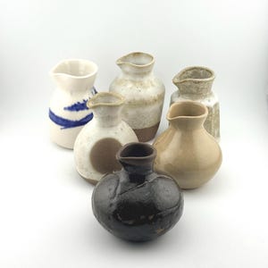 May include: A collection of six small ceramic pitchers with varied finishes. One is white with blue brushstrokes, another is speckled cream, one has a brown circle, one is textured grey, one is smooth tan, and one is a dark, glossy brown.