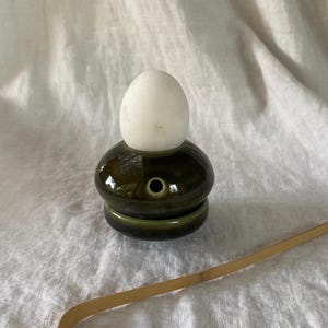 May include: A dark green ceramic tea ceremony accessory with a white egg-shaped object on top. A bamboo whisk lies to the side. The ceramic piece has a small hole and a circular base. The background is a white, textured fabric.