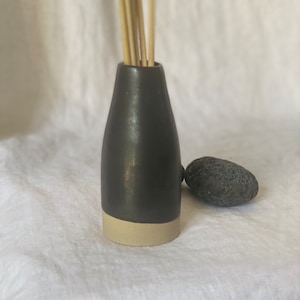 Op de afbeelding: Een zwarte keramische diffuserfles met een beige rand, gevuld met houten diffuserstokjes. De fles staat op een wit linnen oppervlak.