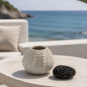 Peut inclure: Un vase en céramique blanc cassé de forme ronde avec une surface texturée et épineuse rappelant une coquille d'oursin, placé à côté d'une petite roche volcanique noire poreuse. Le vase présente une large ouverture circulaire au sommet.