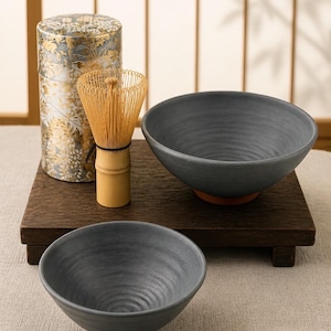 Peut inclure: Un ensemble de deux bols en céramique gris foncé avec un intérieur texturé et une base brun rougeâtre, accompagné d'un fouet en bambou clair et d'un récipient cylindrique à motif floral doré et argenté sur fond gris.