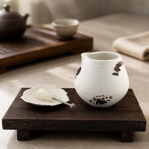 Peut inclure: Une crémière en céramique blanche avec des motifs floraux noirs et bruns est posée sur un plateau en bois foncé. Une petite coupelle blanche avec du sucre en roches et une cuillère en bois sont également sur le plateau. Thème : service à thé.