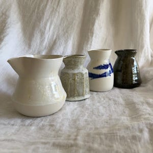 Può includere: Quattro piccoli vasi in ceramica in fila, ognuno con una forma e finitura uniche. Il primo è bianco sporco con la parte superiore svasata, il secondo è strutturato con una tonalità verde chiaro, il terzo è bianco con pennellate blu e l'ultimo è marrone scuro.
