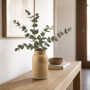 Op de afbeelding: Een cilindrische, mat-beige keramische vaas met verschillende stelen van groene eucalyptusbladeren. De vaas heeft een eenvoudige, afgeronde rand en een gladde, gelijkmatige textuur, wat een neutrale basis vormt voor het blad.