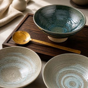 Peut inclure: Deux bols en céramique beige avec des motifs en spirale bleu clair, un bol en céramique émaillée vert foncé avec une finition texturée et une cuillère en laiton à long manche avec un cuilleron circulaire, disposés sur un plateau rectangulaire.