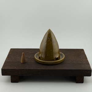 Puede incluir: Un porta incienso de madera marrón oscuro con un pequeño cono de incienso marrón. El porta incienso es de cerámica verde oliva oscuro en forma de cono con una base de platillo a juego. El porta incienso se encuentra sobre una plataforma de madera marrón oscuro.