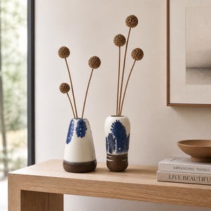 May include: Two ceramic vases with a white base, dark blue brushstroke patterns, and brown glazed bottoms. Each vase holds dried brown spherical flower stems. The vases are positioned on a wooden surface next to stacked books and a wooden bowl.