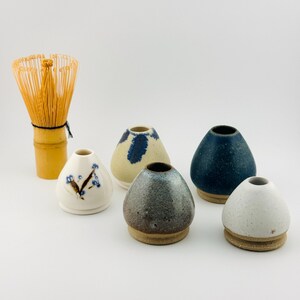 Peut inclure: Un fouet à matcha en bambou et cinq petits vases en céramique aux glaçures variées. Les vases comprennent un modèle blanc à motifs floraux bleus, un crème à taches bleues, un bleu foncé moucheté, un gris métallique et un blanc moucheté.