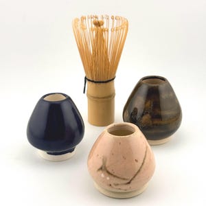 Peut inclure: Un fouet à matcha en bambou est placé derrière trois petits récipients en céramique. L'un est bleu foncé brillant, un autre est rose clair tacheté de lignes sombres, et le troisième est brun foncé marbré. Chaque récipient est arrondi.