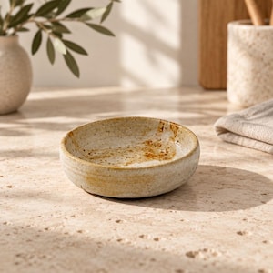 Op de afbeelding: Een kleine, ronde en ondiepe keramische kom met een gespikkelde beige en lichtbruine afwerking. De binnenzijde heeft een textuur met bruine en goudbruine vlekken, terwijl de buitenzijde een glad uiterlijk heeft met subtiele ribbels.