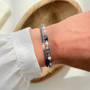 Könnte beinhalten: Zwei Armbänder auf einem Handgelenk. Eines ist ein dunkelblaues Perlenarmband, das andere ein Armband mit weißen Perlen und blauen und grauen Steinen. Die Armbänder befinden sich an einem Handgelenk mit einem weißen Langarmhemd.