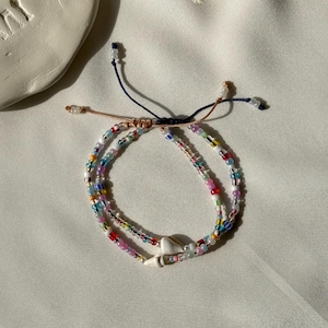 Könnte beinhalten: Ein handgefertigtes Perlenarmband mit einem Muschelanhänger. Das Armband hat eine Vielzahl von bunten Perlen in Rosa-, Blau-, Gelb- und Grüntönen. Das Armband ist mit einer marineblauen und beigefarbenen Schnur gebunden.