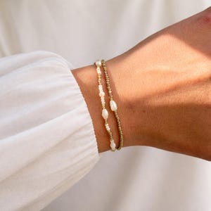 Könnte beinhalten: Zwei zarte Armbänder. Eines hat kleine goldene Perlen, das andere weiße Perlen und kleine goldene Perlen. Die Armbänder werden an einem Handgelenk vor weißem Hintergrund getragen.